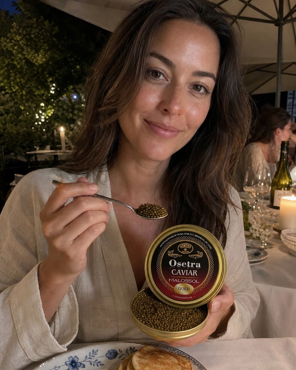 A woman at a candlelit dinner holds a spoonful of caviar over a tin labeled 'Osetra Caviar Malossol Gold' while smiling at the camera.
