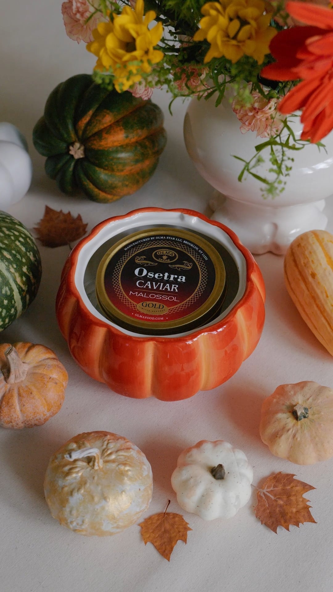 Orange pumpkin-shaped container opened to reveal an Ossetra caviar tin, surrounded by decorative gourds and fall leaves.